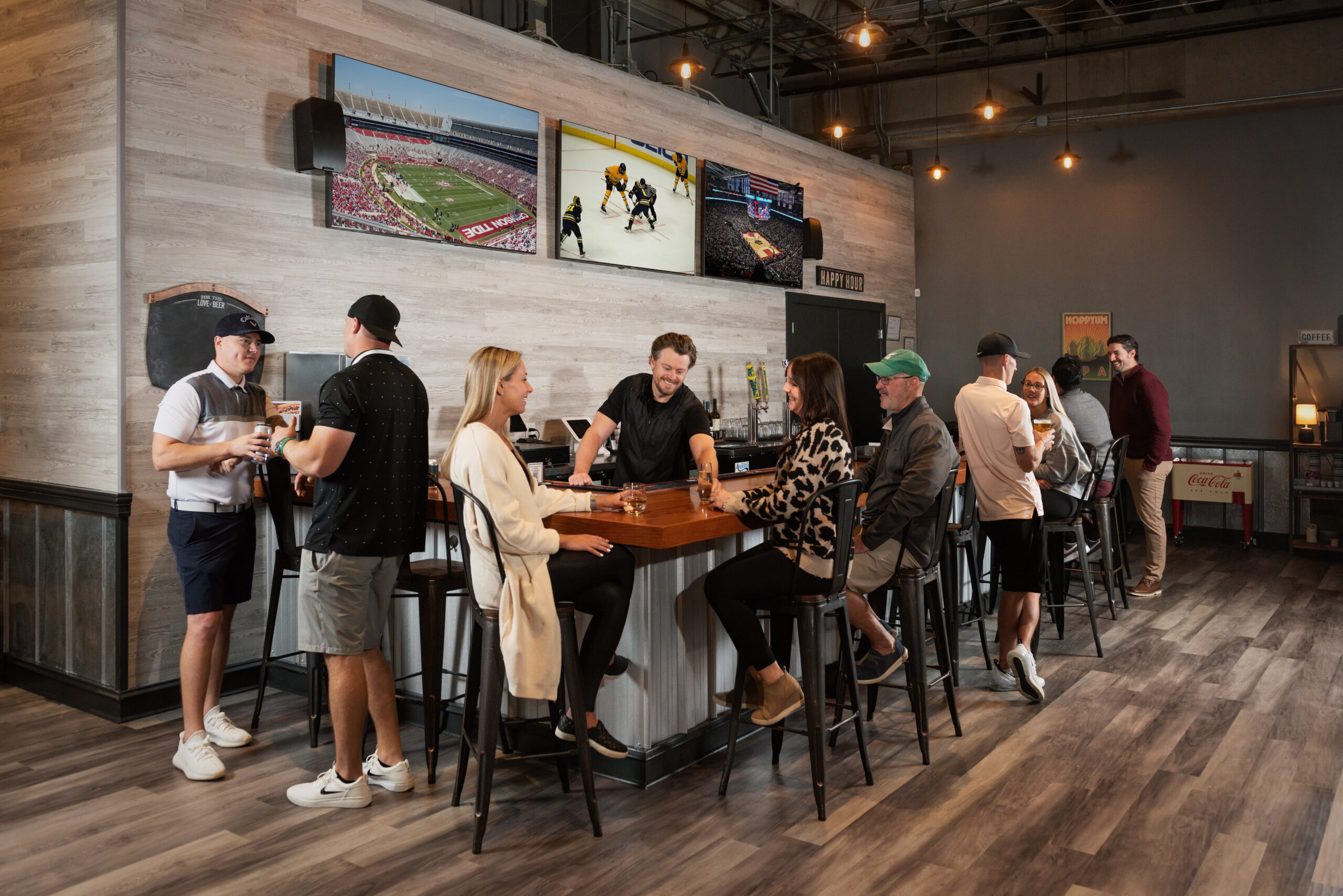 People socialize at a long wooden bar counter, drinks in hand, with large sports screens on the wall showing games in a casual venue.