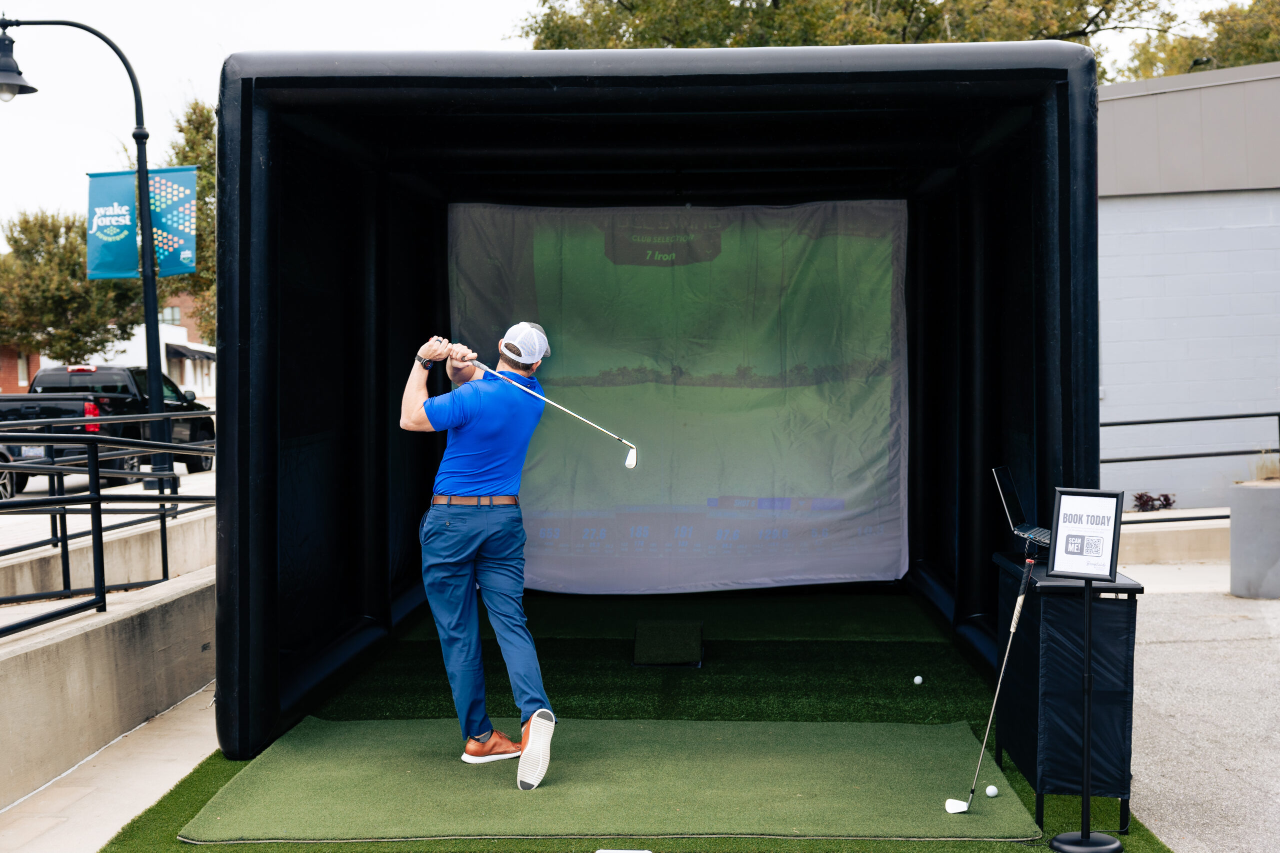 Golfer in a blue shirt and cap swinging a club inside a black outdoor golf simulator booth with a green screen behind him and a Wake Forest banner nearby.”