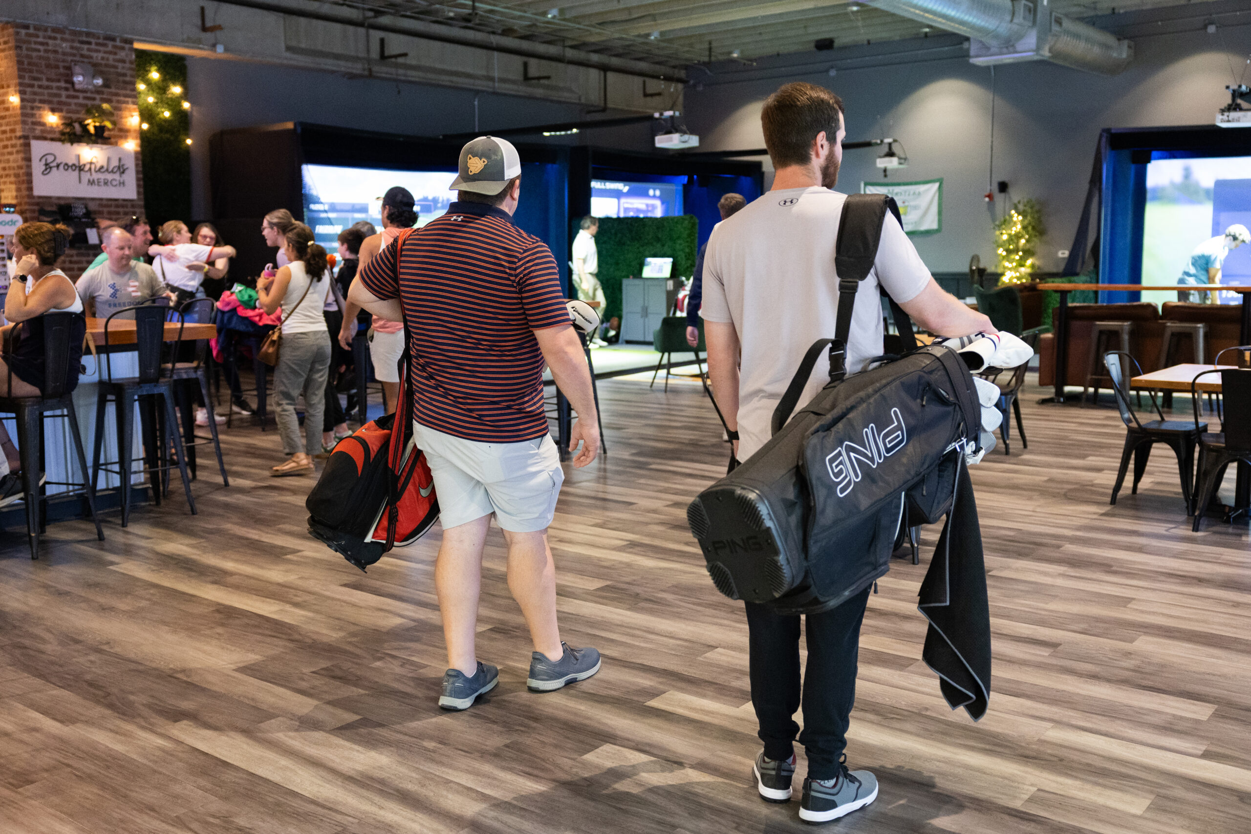 Two men carry large golf bags on their backs, walking through a busy indoor sports venue with seating and people in the background.